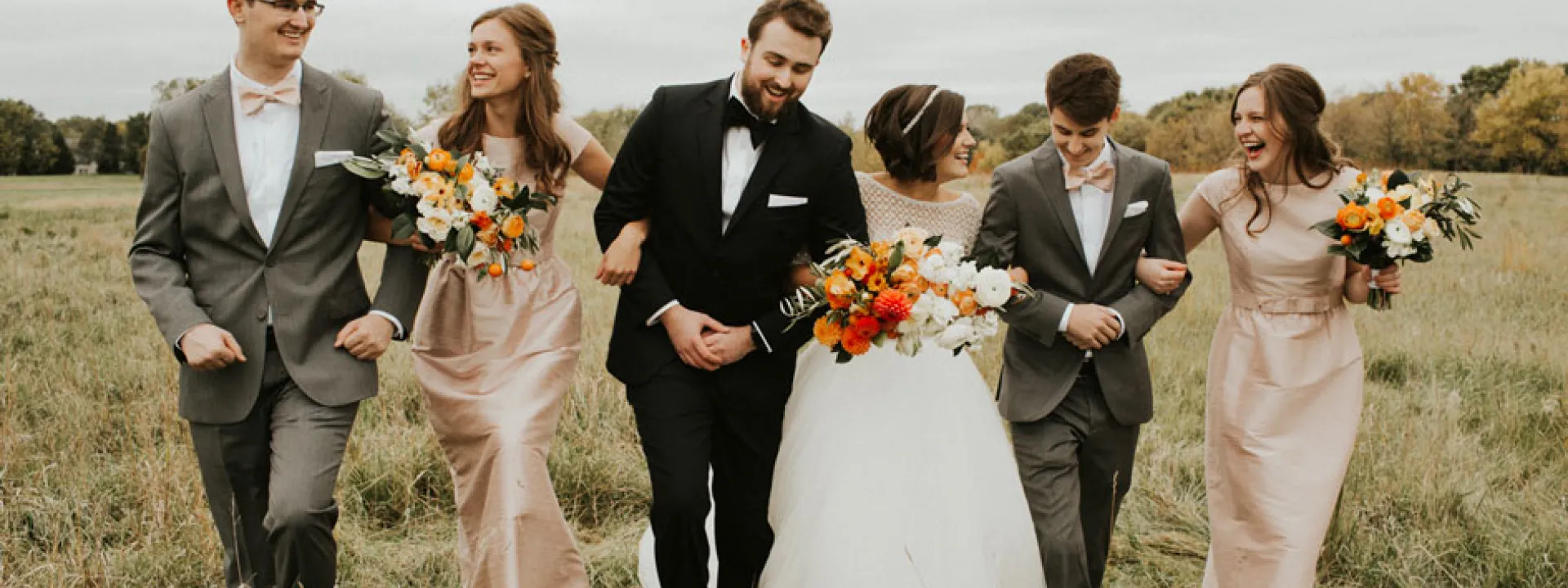 The bridal party, in shades of orange and pastel pink, walks through a rolling field