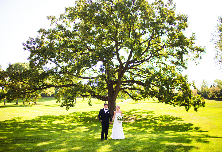 Jake and Paige hold hands in front of a tree during their wedding at the Wayzata Country Club,
