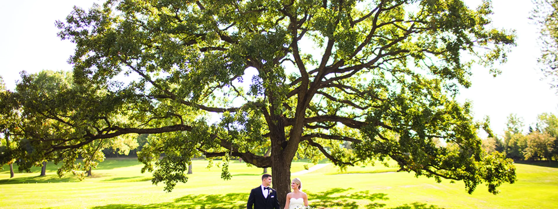 Jake and Paige hold hands in front of a tree during their wedding at the Wayzata Country Club,